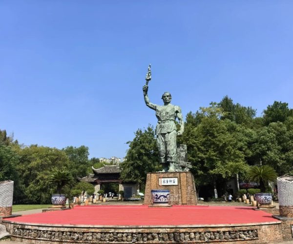 Ancient Kiln Folk Customs Museum,Jingdezhen,China tour Bronze statue of a man holding a raised torch on a brick pedestal, surrounded by a circular red platform and trees in a park.