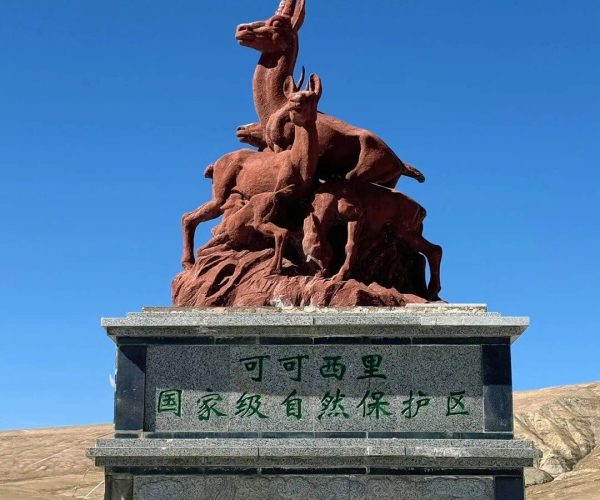 Hoh Xil Nature Reserve,Golmud,China tour Red sculptural group of gazelles perched on a rocky pedestal, with a leaping gazelle at the top, set against a clear blue sky and a stone plinth bearing Chinese text for a national nature reserve.