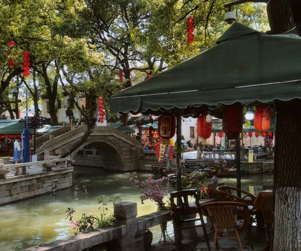 Tongli Water Town,Suzhou,Chinatravel Outdoor cafe beside a canal with a stone bridge, red lanterns, and shaded tables under green awnings.