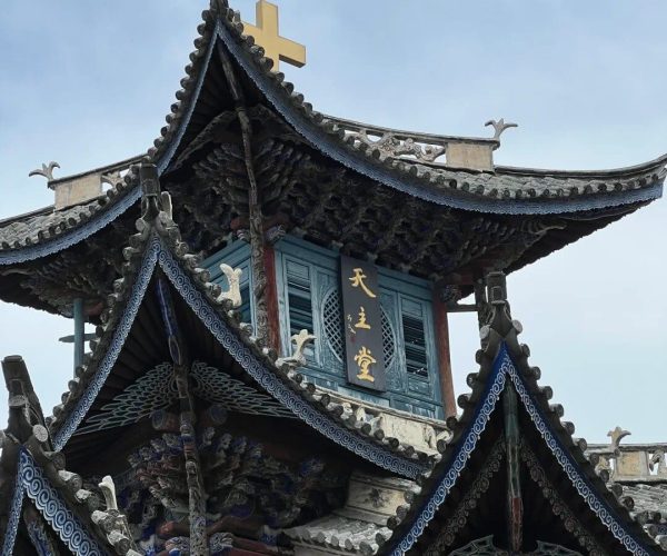 Xizhou & Zhoucheng Bai Villages,Dali,Chinatravel Ornate multi-tiered Chinese temple roof with decorative blue eaves and a gold cross atop the central peak against a blue sky.
