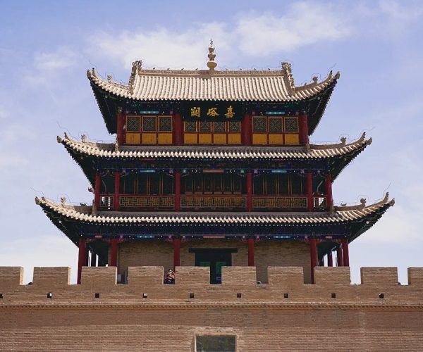 Jiayuguan Pass,Jiayuguan,China tour Three-tier Chinese pagoda atop a historic brick city wall with an arched gate; silhouetted visitors pass through the arch against a blue sky.
