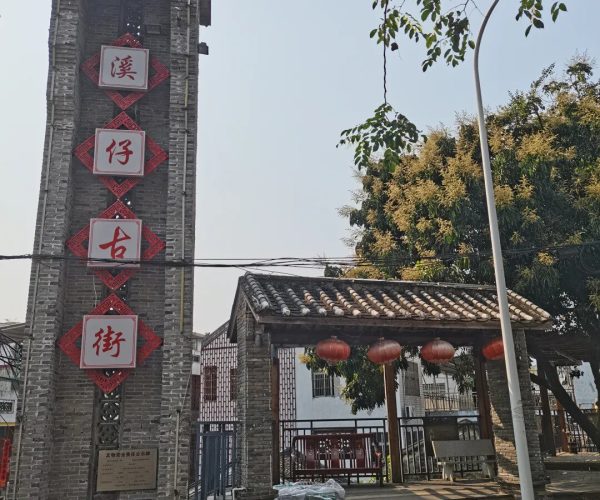 Jiaji Old Town,Qionghai,Chinatravel Brick gateway pillar with vertical red plaques bearing Chinese characters, opening onto a traditional street with hanging red lanterns and a tiled roof structure to the right.