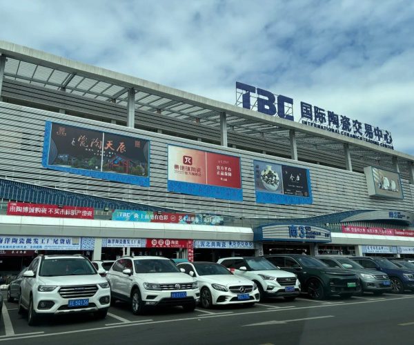 International Ceramics Trade Center,Jingdezhen,China tour Facade of the International Ceramics Trade Center (IBTC) with blue signage and large ad banners along the front, row of parked cars in the foreground.