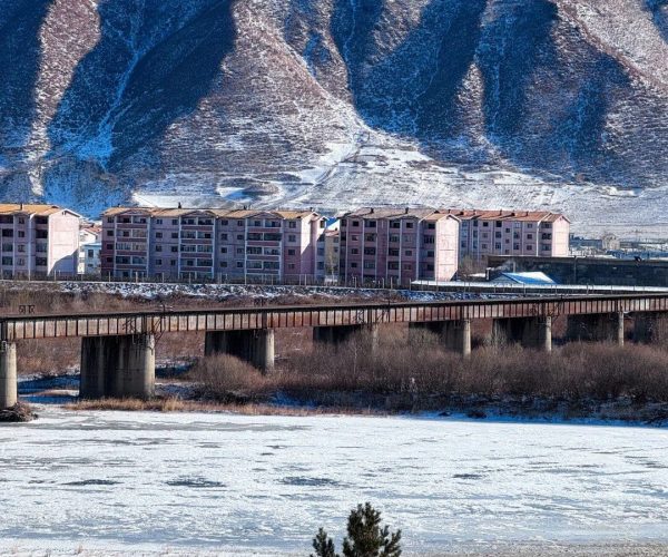 Tumen Por,Yanbian,China tour Snowy mountain range behind a row of pink apartment buildings and a rust-colored bridge over a frozen river.