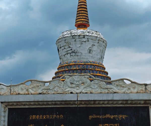 Ta’er Monastery,Xining,Chinatrip Weathered white stupa with a tall striped orange and black spire, set against a cloudy blue sky; ornate carved base and colorful decorative bands near the pedestal.