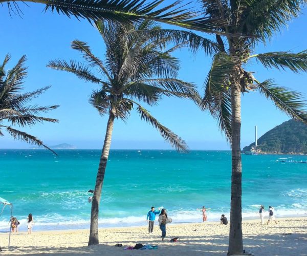 Dadonghai Beach Area,Sanya,Chinatravel Sunny tropical beach scene with palm trees, turquoise water, and people on the sandy shore under a clear blue sky.