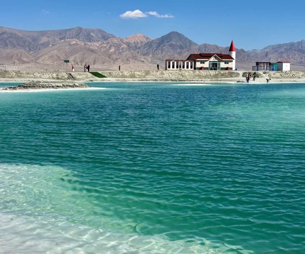 Dachaidan Emerald Lake,Haixi Prefecture,China tour Turquoise lake with a white-sand shore, distant red-roofed buildings, and arid mountains under a clear blue sky.