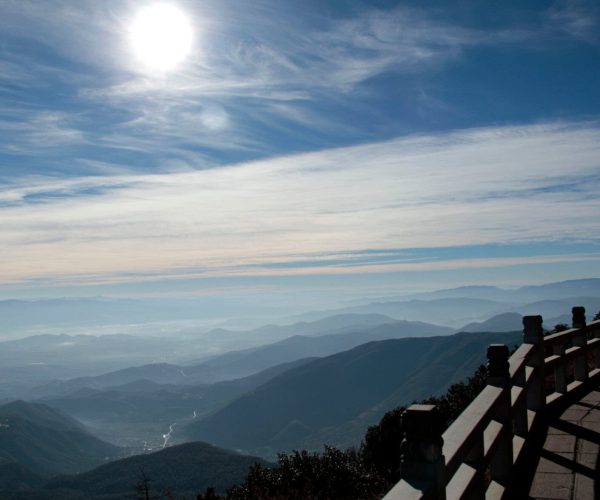 Dali Weather,Dali,Chinatravel Mountain range under a bright sun with wispy clouds and a wooden railing in the foreground.