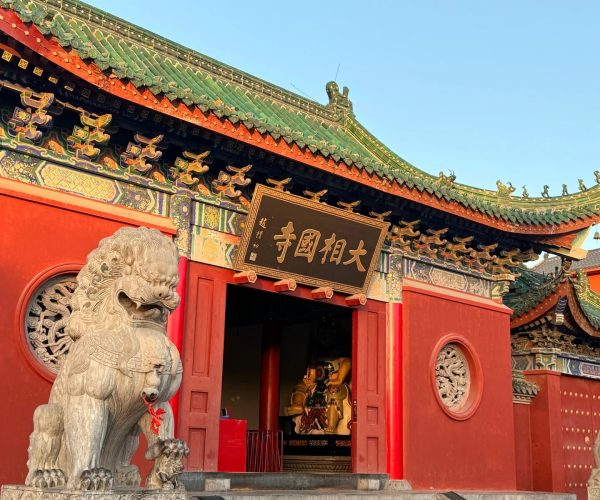 Daxiangguo Temple,Kaifeng,Chinatravel Red temple entrance with a stone guardian lion in the foreground and a black plaque with gold Chinese characters above the doorway, ornate colorful eaves above.