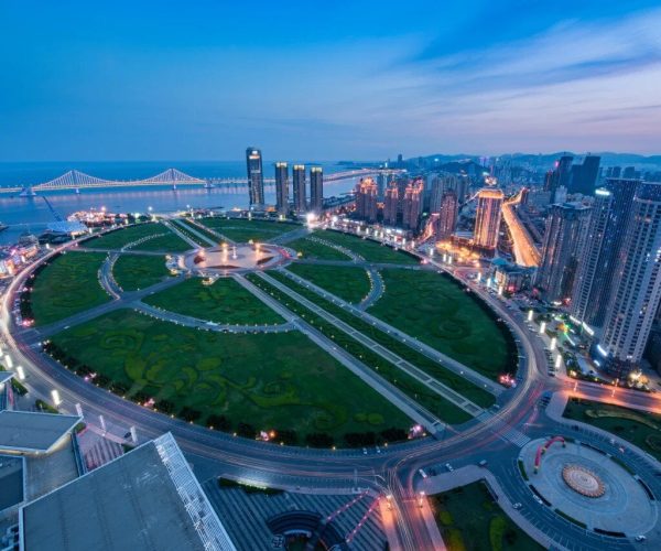 Xinghai Square,Dalian,Chinatravel Aerial view of a circular city park with radiating roads and a skyline of high-rise buildings along the waterfront at dusk.