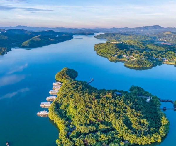 Tianmu Lake,Changzhou,China tour Aerial view of a lush, tree-covered island in a calm blue lake with a row of white buildings along the shore and distant green hills in the background, under a clear sky.