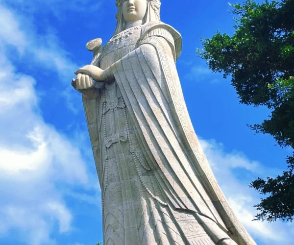 Meizhou Island Mazu Temple,Putian,Chinatravel Tall stone statue of a standing bodhisattva in ornate robes, holding a small round object, against a bright blue sky.
