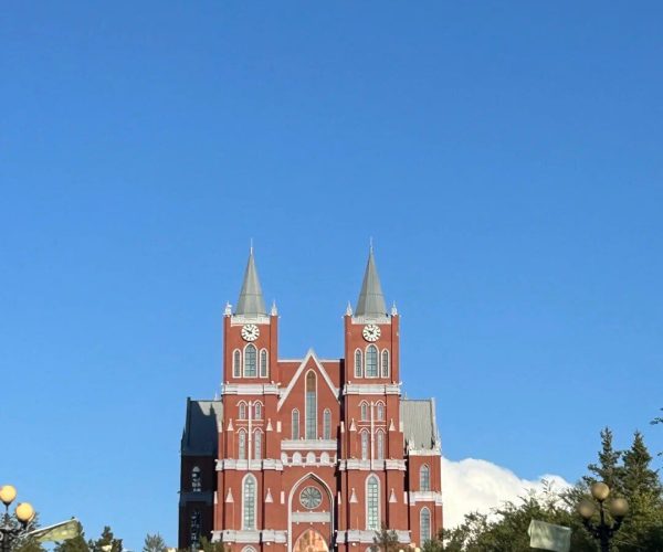 Wedding Palace,Manzhouli,China tour Red brick cathedral with twin spires at the end of a straight path lined with lamp posts and trees under a blue sky.
