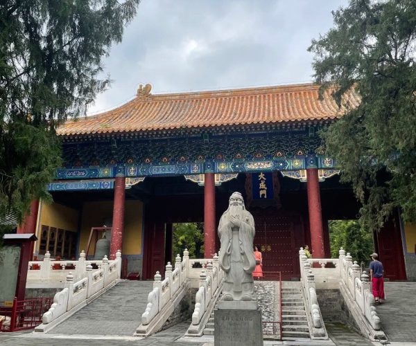 Confucius Temple,Qufu,China tour Stone statue of a wise scholar on a pedestal in a temple courtyard, with white stone railings and gray paving in front of a traditional Chinese temple.