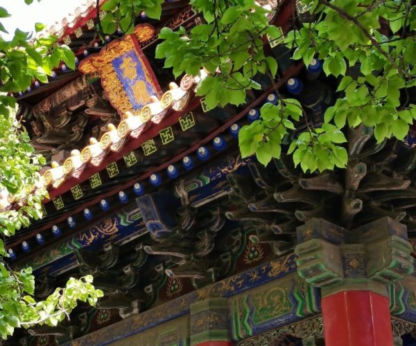 Confucius Mansion,Qufu,China tour Colorful traditional Chinese temple roof with red columns and intricate blue-green carvings partially shaded by green leaves.