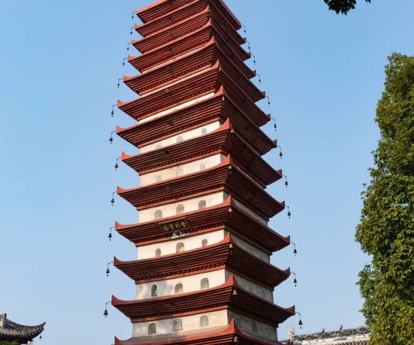 Baoguang Temple,Leshan,Chinatravel Tall multi-tiered Chinese pagoda with red eaves and hanging bells, set beside temple buildings under a blue sky.