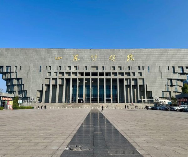 Shandong Museum,Jinan,China tour Large gray modern building with gold Chinese characters on top, set in a wide, paved plaza under a clear blue sky; people walk toward the entrance.