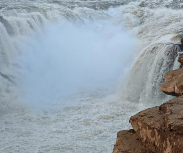 Hukou Waterfall on Yellow River,Linfen,Chinatravel Massive waterfall with mist, rugged rocks in the foreground and distant hills with traditional buildings along the shore.