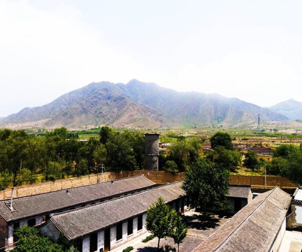 Shanxi Weather,Shanxi,Chinatravel Mountain range rises behind a village with tiled-roof buildings in the foreground and green trees.