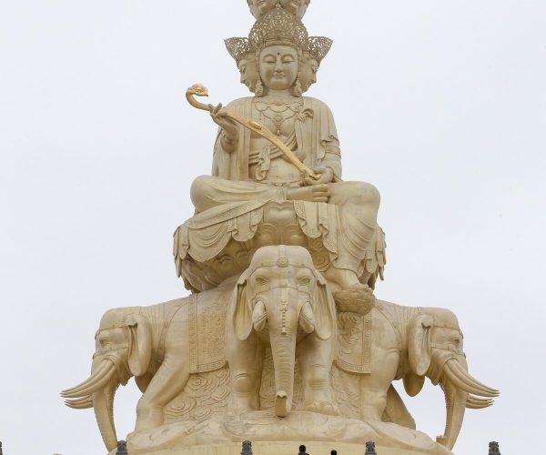 Mount Emei Scenic Area,Leshan,Chinatravel Large beige statue of a multi-headed deity seated on an elephant throne, atop a stepped platform, holding a curved staff against a pale sky.