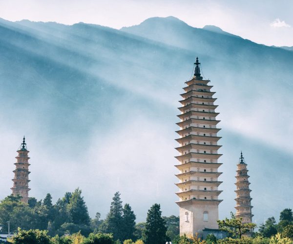 Dali Three Pagodas,Dali,Chinatravel Three tall, multi-tiered pagodas rise in a garden with trees, mountains, and a hazy blue sky behind them, creating a serene landscape.