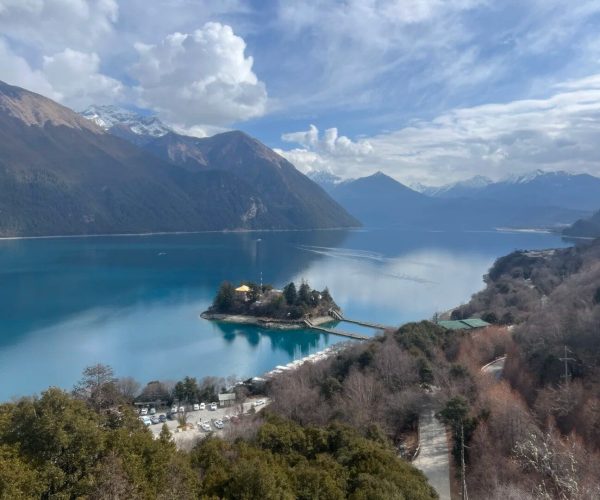 Basong Lake,Nyingchi,China tour Turquoise lake surrounded by snow-capped mountains with a small island containing a yellow-roofed building connected by a pier/boardwalk.