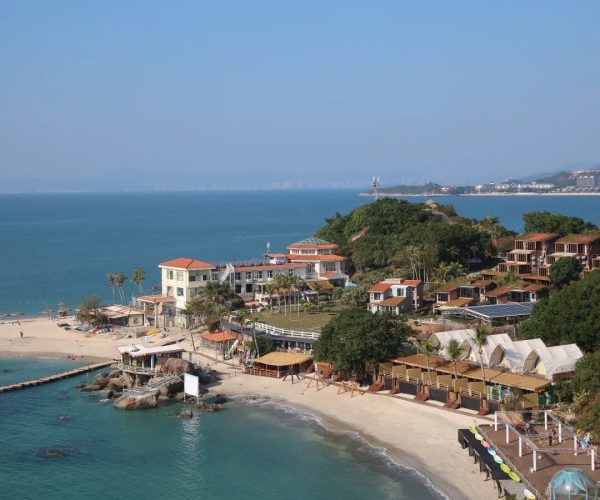 Xunliao Bay,Huizhou,Chinatravel Aerial view of a seaside resort with white buildings and orange roofs along a sandy bay, turquoise water, and palm trees.