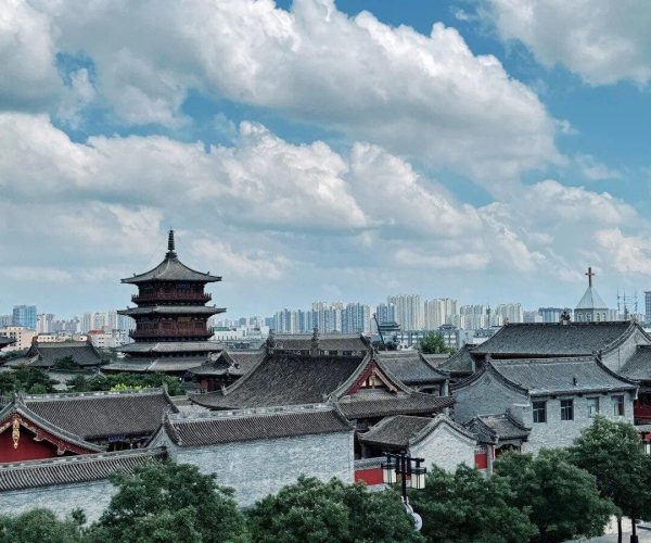 Pingcheng Ancient Town,Datong,Chinatravel Traditional Chinese temple complex with a multi-tier pagoda against a blue sky, row of grey-tiled roofs, and urban skyline in the distance behind green trees.