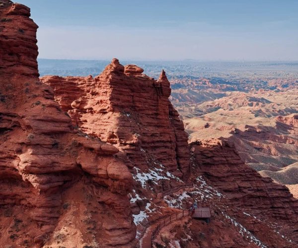 Pingshanhu Grand Canyon Red sandstone cliffs and a winding wooden walkway along a steep canyon, with snow patches and a distant blue horizon under a clear sky.