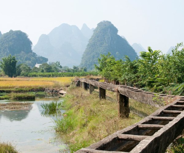 Guangxi Weather,Guangxi,Chinatravel Rural scene with a long weathered wooden irrigation trough beside fields and distant karst mountains under a pale sky.