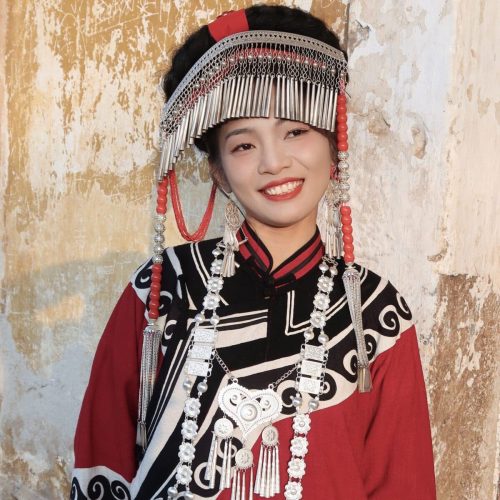 Yi ethnic costumes,Yunnan,HanfuYoung woman in a red and black traditional dress with a large silver headdress and layered silver jewelry, smiling against a textured wall.