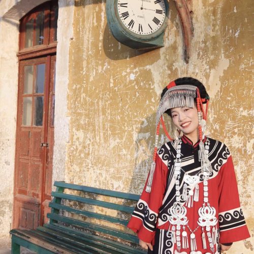 Woman in ornate red, black, and white traditional dress with silver headdress Yi ethnic costumes,Yunnan,Hanfu standing beside a green bench under a vintage clock on a peeling wall.