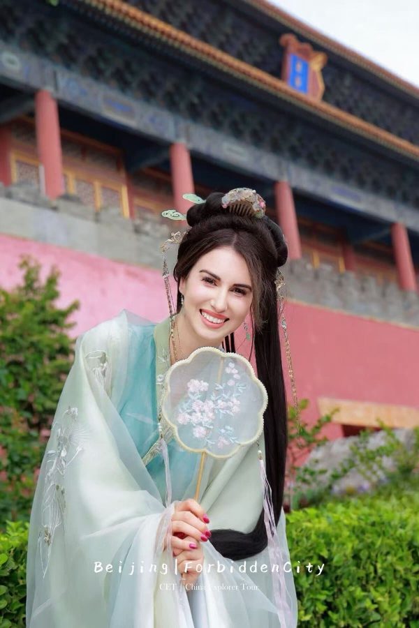 hanfu photoshoot, the forbidden city, beijing, female, solo, chinatravel, Smiling woman in pale blue traditional Hanfu holding a lace fan, posing outdoors near a temple/ Forbidden City backdrop.
