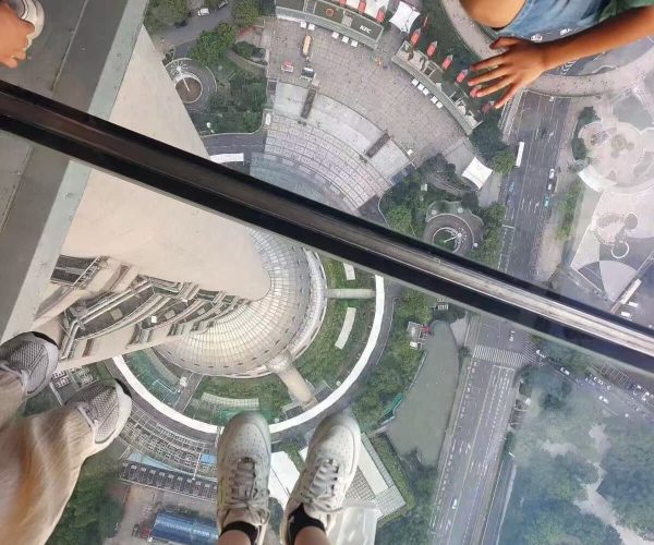 Pearl Tower,Shanghai,Chinatour People stand on a transparent glass floor high above a city, looking down at streets and buildings below, with their feet visible at the edge.