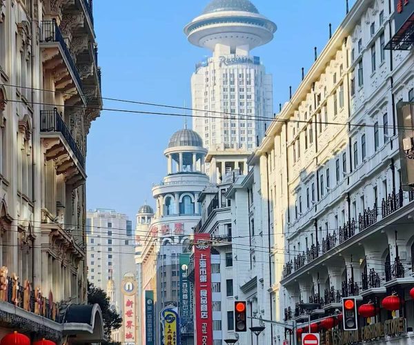 Nanjing Road,Chinatour Urban street in Shanghai with colonial-style buildings, red lanterns, and colorful banners; the Oriental Pearl Tower rises in the distance against a clear blue sky.