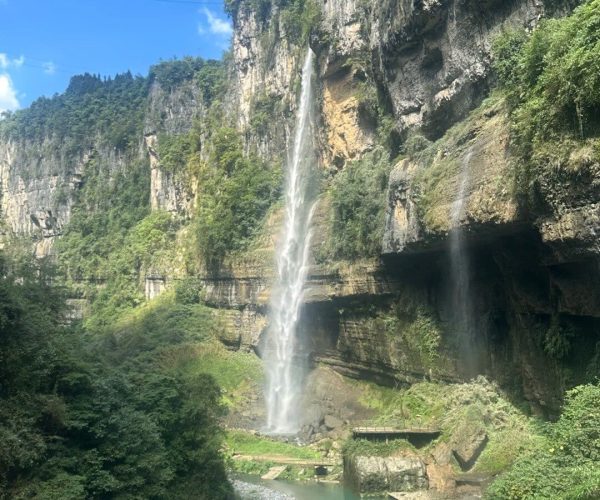 Enshi Grand Canyon,Enshi,Chinatravel Tall waterfall plunging from a rocky cliff into a narrow pool below, surrounded by lush green vegetation and a bright blue sky.