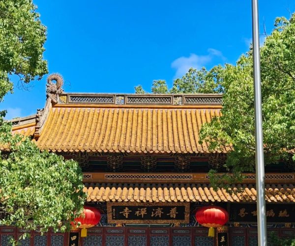 Huiji Temple,Zhoushan,Chinatravel Traditional Chinese temple with orange-tiled roof, red lattice walls, gold calligraphy plaques, red lanterns, and visitors outside on a sunny day.