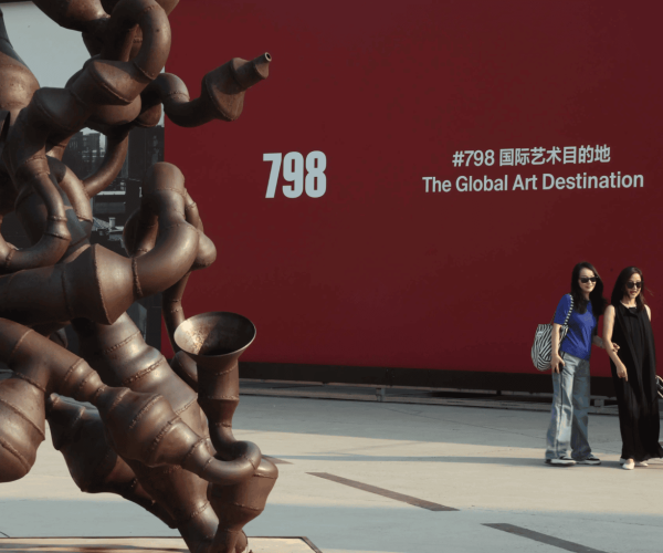 798, beijing, chinatravel, Rusty abstract sculpture on the left with two women and a white dog walking by a red wall that reads '798 The Global Art Destination' on the right.