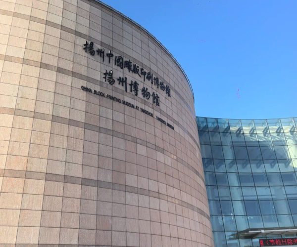 Yangzhou Museum,Yangzhou,China tour Curved beige stone wall of a museum with Chinese and English text, glass entrance to the right under a blue sky.