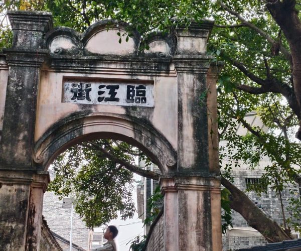Yangmei Ancient Town,Nanning,Chinatravel Old stone archway with a faded Chinese inscription, set in a tree-lined street; people and scooters pass beneath.