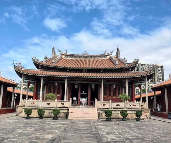 Jieyang Confucian Temple,Jieyang,Chinatravel Front view of a traditional Chinese temple with tiered tiled roof and red wooden pillars, set in a stone-paved courtyard with potted shrubs and a central incense burner; blue sky above with modern buildings in the distance.