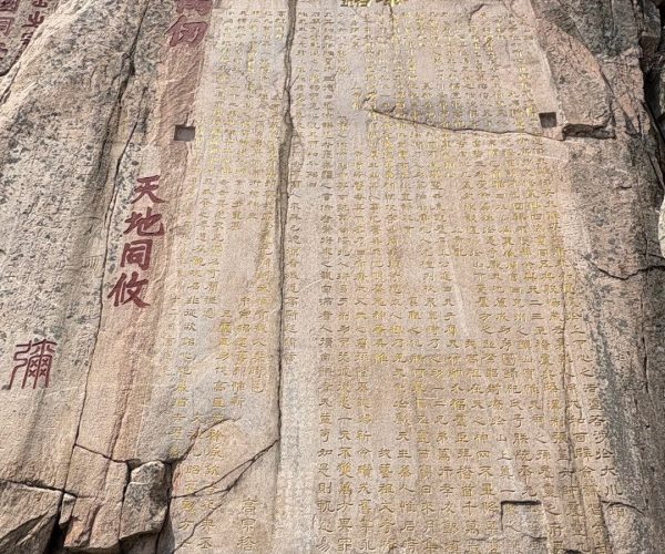 Cliff Inscriptions,Tai'an,China tour Large granite rock face covered in vertical Chinese inscriptions in gold and red paint, with a small cave opening at the bottom left and barren trees visible above.