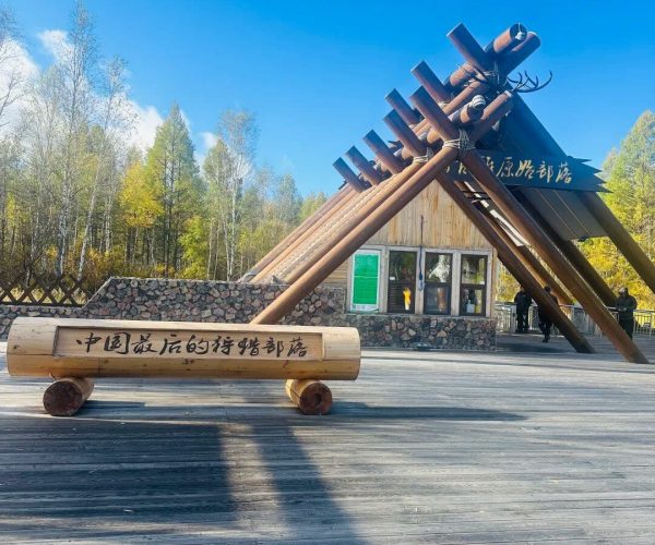 Aoluguya Reindeer Tribe,Hulunbuir,Chinatrip Outdoor wooden sculpture on a plaza: a triangular arch formed by thick logs, with a bench inscribed in Chinese and a small building in the background among trees on a sunny day.