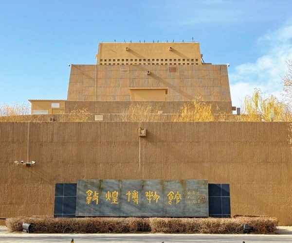 Dunhuang Museum,Dunhuang,Chinatrip Exterior view of a beige museum with a dark stone sign bearing gold Chinese characters, low hedges, and a paved plaza with cones against a blue sky.