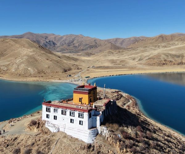 Nichiren Temple,Shannan,China tour A small white and orange monastery perched on a rocky peninsula beside a bright blue lake, with arid hills and mountains in the background under a clear sky.