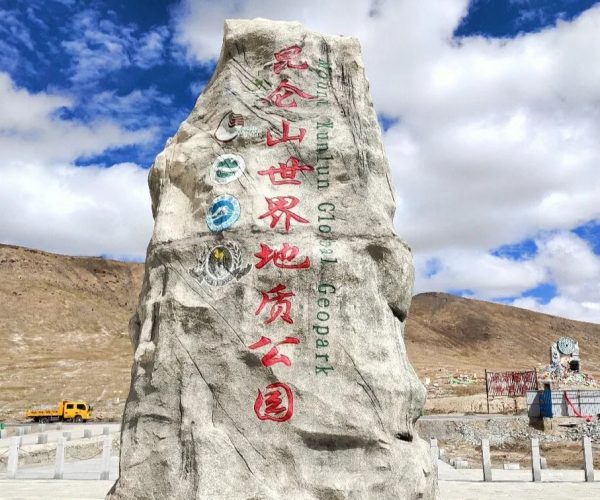 Kunlun Mountains Geopark,Golmud,China tour Large rock with red Chinese inscriptions and vertical 'Global Geopark' text, set in a desert landscape under a blue sky with hills in the background; a yellow truck is visible nearby.