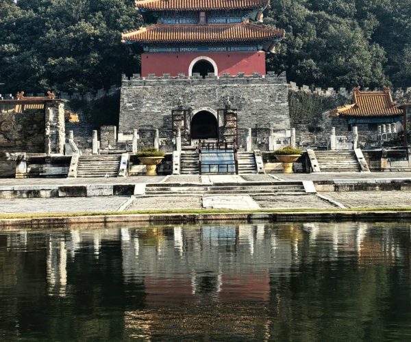 Mingling Mausoleum,Yichang,Chinatravel Traditional Chinese pavilion with orange-tiled roofs standing on a stone fortress beside a calm reflecting pool, with trees in the background and its reflection in the water.”] }**Wait**: Need multiple options. I'll correct. Let's provide 5.