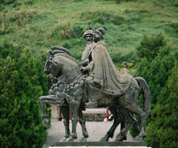 Zhaojun Tomb,Hohhot,Chinatrip Bronze statue of two armored riders on galloping horses, mounted on a stone pedestal in a park with a green hillside behind.