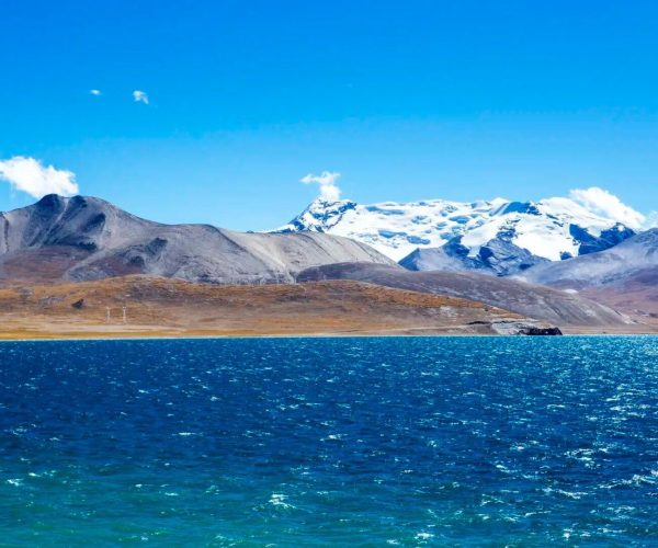 Pumoyong Lake,Shannan,China tour Snowy mountains rise behind a brown, barren hillside and a bright blue sky, with a deep turquoise lake in the foreground.