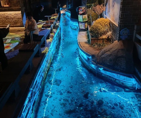 Qushuiting Street,Jinan,China tour Nighttime narrow canal lined with blue-lit water, outdoor seating on the left and a mural wall on the right.